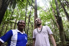 Foto mostra o pesquisador, de camiseta branca e colares indígenas, ao lado do indígena Pedro Baniwa, de camiseta azul, no meio da floresta